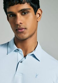Hellblaues Polohemd mit klassischem Kragen, versehen mit dezentem gesticktem Logo auf der Brust und zwei Knopfverschlüssen. Glatte Stoffoberfläche.