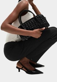 Czarna, teksturowana torebka z podwójnymi uchwytami, o zdefiniowanym kształcie i błyszczącym wykończeniu, trzymana w tle jasnego topu i czarnych spodni.