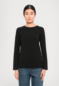 Moteris vilkinti juodą ilgomis rankovėmis Armani Exchange marškinį su logotipais ant krūtinės ir mėlynomis džinsomis, stovinti prieš paprastą šviesų foną.