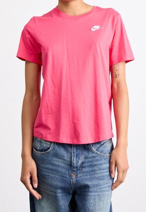 Persoon die een roze Nike t-shirt en blauwe spijkerbroek draagt met een kleine teddybeer-tatoeage op de linkerarm.