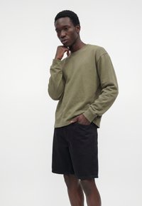 Jeune homme portant une chemise à manches longues vert olive et un short noir, debout avec une main dans la poche et l'autre près du menton.