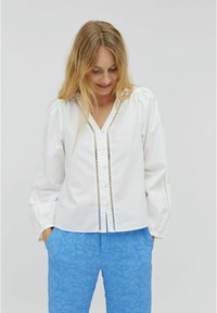 White button-up shirt with puff sleeves, featuring lace detailing along the placket. Paired with textured blue pants.