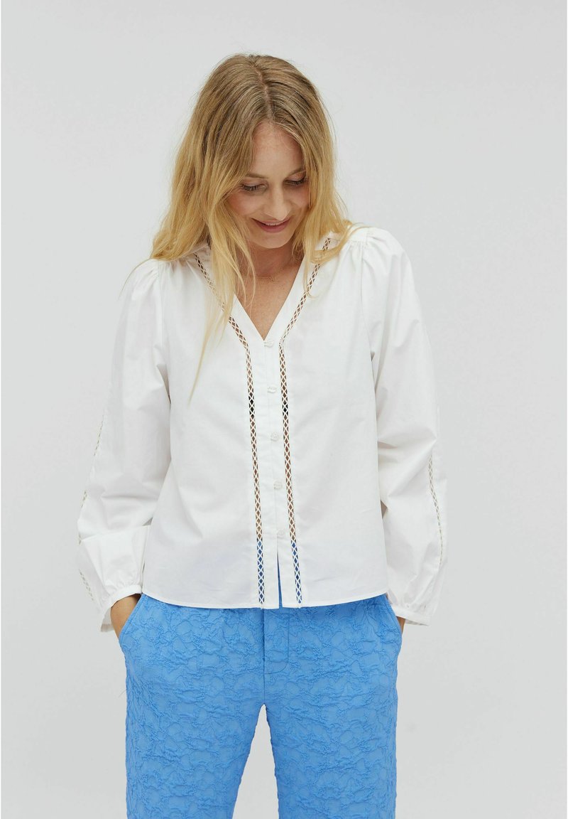 White button-up shirt with puff sleeves, featuring lace detailing along the placket. Paired with textured blue pants.