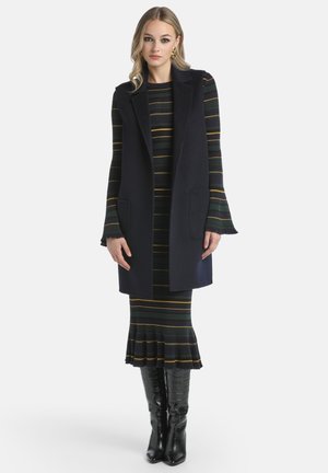 Marinefarbener ärmelloser Weste mit Seitentaschen über einem figurbetonten gestreiften Kleid in Grün-, Gelb- und Schwarztönen mit einem ausgestellten Saum. Schwarze kniehohe Stiefel.