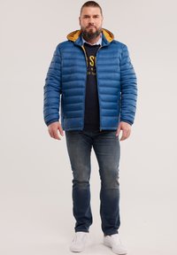 Mann mit Bart, der eine blaue Steppjacke mit gelber Kapuzenfutter, dunkles Sweatshirt, Jeans und weiße Turnschuhe trägt und vor einem einfarbigen Hintergrund steht.
