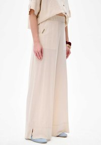 Hellbeige, weite Hosen aus weich leichtem Stoff. Merkmale sind seitliche Taschen, ein flacher Bund und geschlitzte Saums. Kombiniert mit hellblauen Schuhen.