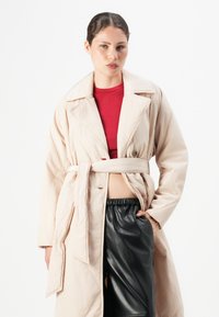 Kvinne ikledd en beige belted frakk over en rød topp og svart skinnskjørt, står med den ene hånden i lommen mot en hvit bakgrunn.