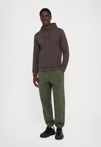 Mann iført en mørkebrun hettegenser og olivengrønne joggebukser med sorte joggesko, stående med en hånd i lommen mot en ensfarget bakgrunn.