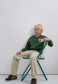 Sudadera verde con gráfico blanco, pantalones beige y zapatillas marrón oscuro con detalles en amarillo. Sentado en una silla plegable verde.