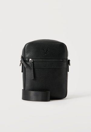 Petit sac bandoulière en cuir noir compact avec poche zippée à l'avant et bandoulière réglable sur fond clair uni.