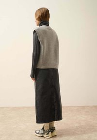 Szary dziany kamizelka na ciemnym golfie, w zestawie z długą czarną spódnicą dżinsową i szarymi butami sportowymi, widziana z tyłu.