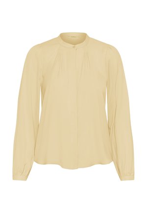Blouse beige à manches longues avec un col rond, des détails plissés près des épaules et des poignets boutonnés sur un fond uni.