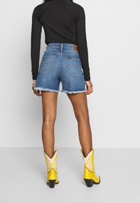 Shorts de denim con un dobladillo deshilachado, combinados con una blusa negra de manga larga y botas vaqueras amarillas con cortes decorativos y un tacón cuadrado.