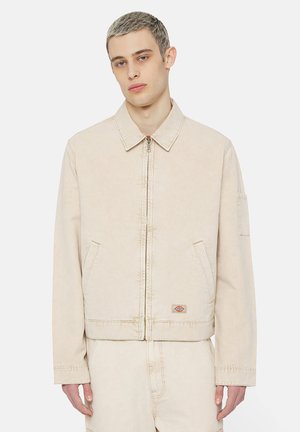Giacca di jeans beige con collo, zip frontale, due tasche laterali e un logo a patch sul lato sinistro. Tessuto morbido, vestibilità comoda.