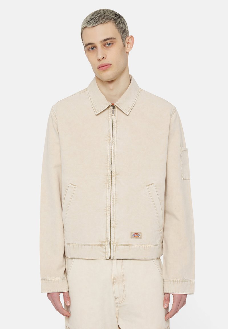 Giacca di jeans beige con collo, zip frontale, due tasche laterali e un logo a patch sul lato sinistro. Tessuto morbido, vestibilità comoda.