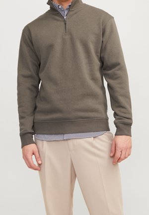 Homme portant un sweat-shirt à fermeture éclair partielle vert olive sur une chemise à rayures boutonnée, avec un pantalon beige, debout devant un fond uni.