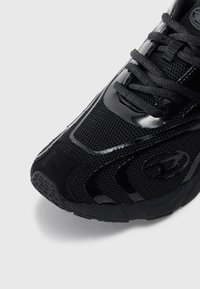 Schwarzer Sportschuh mit einer Kombination aus Mesh- und glatten Synthetikmaterialien, einer strukturierten Sohle und dezentem Branding an der Seite.
