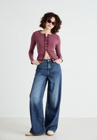 Ribbed burgundy cardigan with button closure, paired with wide-leg blue jeans; model wearing slim, rectangular sunglasses and white sneakers.