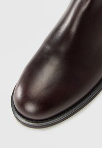 Close-up of smooth, dark brown leather boot toe with black sole and detailed stitching on light background.