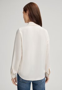 Blusa a maniche lunghe color crema con una texture liscia e polsini con bottoni, dotata di un colletto classico e di un orlo curvo, indossata con jeans blu.