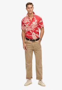 Camisa corta roja con estampado floral blanco, combinada con pantalones marrón claro y zapatillas color crema. Cinturón negro y reloj de pulsera visibles.