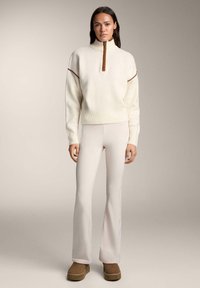 Kremasti pulover s polu-zip izrezom i smeđim patentnim zatvaračem, u kombinaciji sa svijetlo bež hlačama širokih nogavica i smeđim platformama.