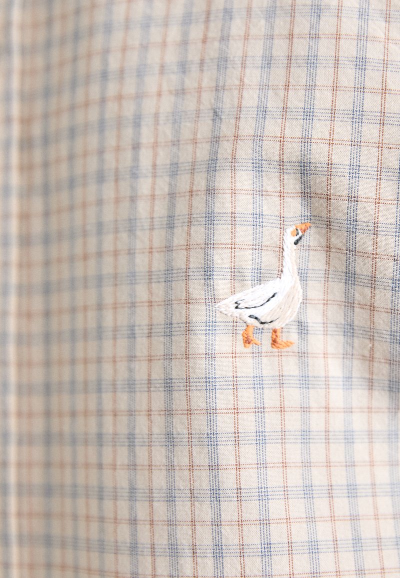 Bomullstyg med ett blått och rött rutmönster, prytt med en detaljerad vit och orange broderad gåsdesign.