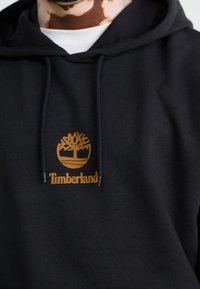 Sudadera negra de tela suave, con un logo dorado de Timberland con un diseño de árbol en el centro, y cordones ajustables en la capucha.