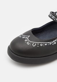 Czarny skórzany but o zaokrąglonym nosku, zdobiony białymi wzorami i paskiem do zapinania. Teksturowana gumowa podeszwa zapewnia przyczepność.