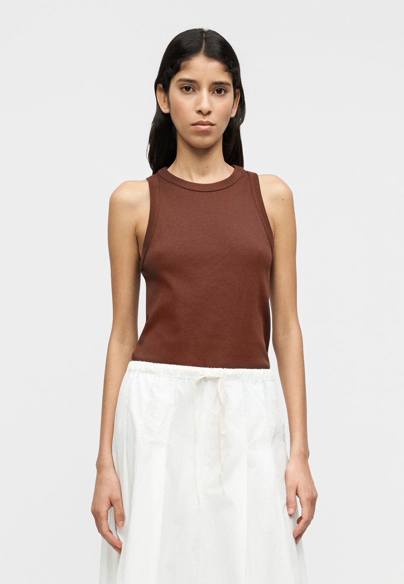 Καφέ ριγωτό τοπ χωρίς μανίκια με στρογγυλή λαιμό κοψη, συνδυασμένο με λευκή φούστα με κορδόνι στη μέση, ουδέτερο φόντο.