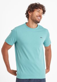 Smiling man with curly hair wearing a light blue short-sleeve t-shirt and navy blue pants, posing with hands behind back.