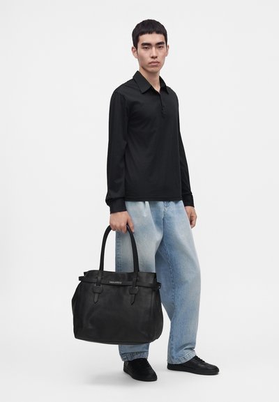 Jeune homme portant une chemise noire à manches longues et un jean bleu clair tenant un grand sac cabas en cuir noir avec deux poignées.