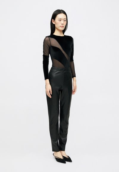 Macaquinho preto com seções transparentes, combinado com calças pretas de couro sintético de cintura alta. Sapatos planos pretos pontiagudos completam o visual.