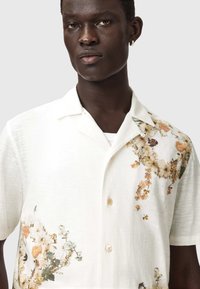 Άνδρας με σκουρόχρωμο δέρμα που φοράει λευκό κοντομάνικο πουκάμισο με μπεζ και καφέ ανθικά μοτίβα στον ώμο και στο κάτω μέρος.