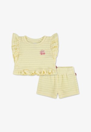 Żółty dwuczęściowy strój z teksturowanym topem z falbanami na rękawach i na dole, w zestawie z dopasowanymi szortami. Charakterystyczny różowy detal z logo.