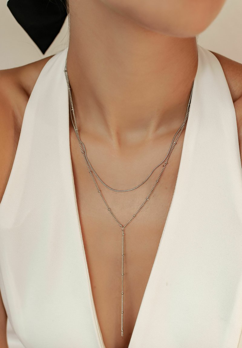 Naszyjniki warstwowe ze srebra o różnych długościach, z gładkimi i teksturowanymi łańcuchami, noszone na białej bluzce z dekoltem w serek.