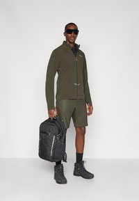 Άνδρας σε πράσινη zip-up ζακέτα και σορτς, φορώντας μαύρες ορειβατικές μπότες και γυαλιά ηλίου, κρατώντας έναν μαύρο σακίδιο σε απλό φόντο.