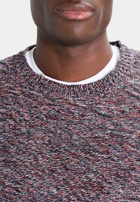 Pull en tricot à col rond avec un motif texturé multicolore comprenant des fils rouges, bleus et blancs, doté d'un col côtelé et porté avec une chemise blanche superposée en dessous.
