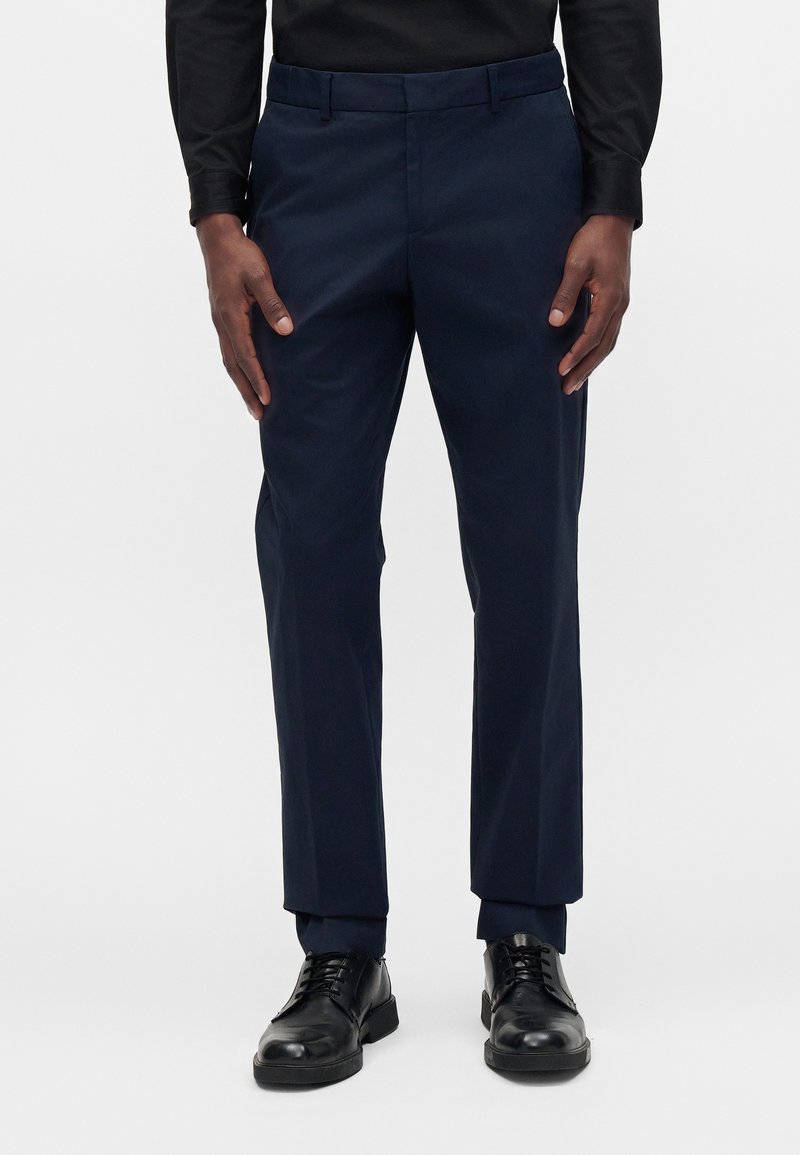 Homme portant un pantalon de costume bleu marine et des chaussures en cuir noir, debout devant un fond uni.