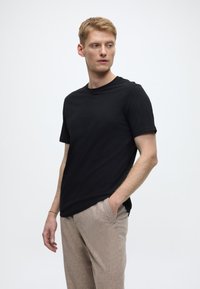 Schwarzes T-Shirt mit kurzen Ärmeln in entspannter Passform und Rundhalsausschnitt, kombiniert mit hellbraunen, strukturierten Hosen. Einfaches Design, minimale Akzente.