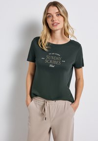 Mladá žena s dlouhými blond vlasy, oblečená v tmavě zeleném tričku s nápisem "Sunday Scribes Club" a béžových kalhotách na šňůrku, má ruce v kapsách.