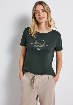 Femme jeune avec de longs cheveux blonds portant un tee-shirt vert foncé avec le texte "Sunday Scribes Club" et un pantalon beige à cordon de serrage, les mains dans les poches.