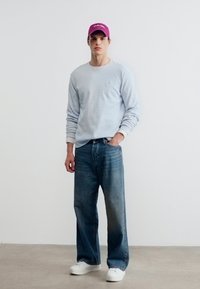 Άνδρας που στέκεται φορώντας ανοιχτό μπλε πουκάμισο με μακριά μανίκια, τζιν με φαρδιά πόδια, λευκά αθλητικά παπούτσια και ροζ καπέλο μπέιζμπολ σε απλό λευκό φόντο.