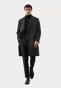 Kabát z vlněné směsi v tmavě šedé barvě s texturovaným povrchem, se zářezovým límcem, bočními kapsami a dlouhým, rovně střiženým střihem, nošený přes černé oblečení.