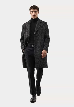 Kabát z vlněné směsi v tmavě šedé barvě s texturovaným povrchem, se zářezovým límcem, bočními kapsami a dlouhým, rovně střiženým střihem, nošený přes černé oblečení.