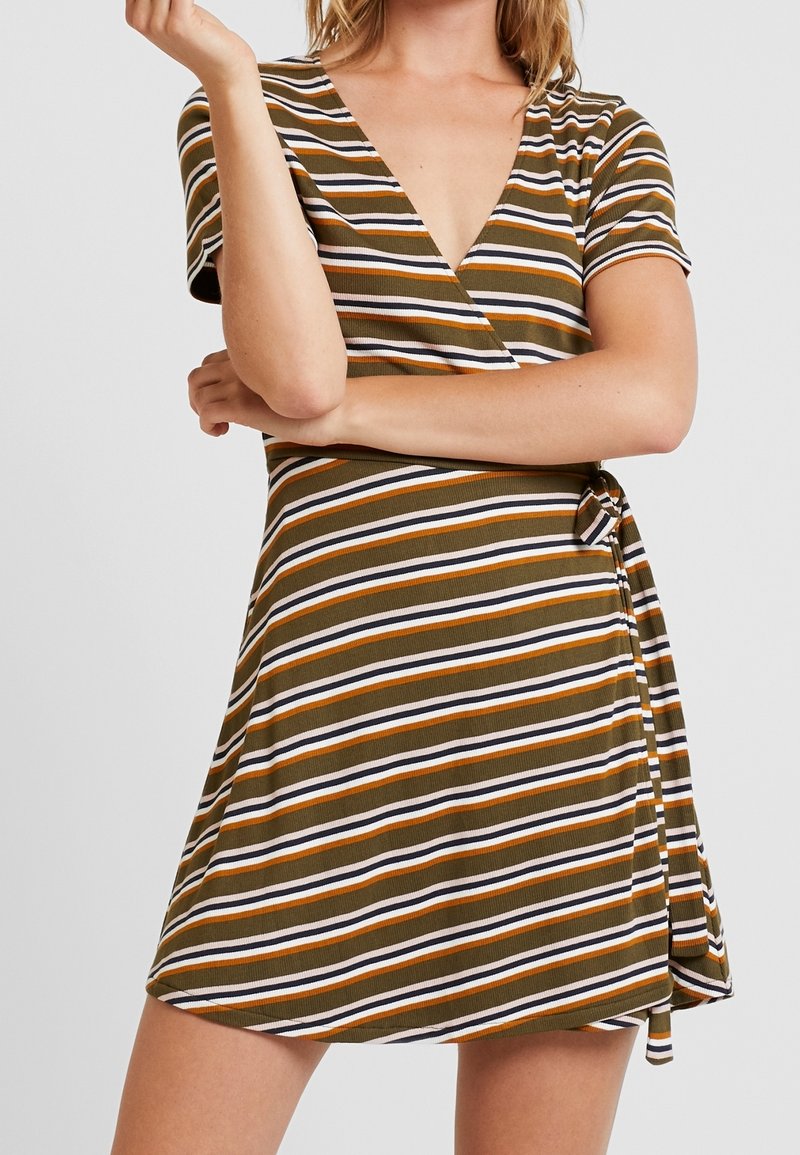 Femme portant une robe portefeuille à manches courtes et col en V avec des rayures horizontales marron, orange, blanc et noir, bras croisés à la taille.