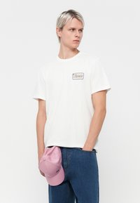 White cotton t-shirt with short sleeves, featuring a small embroidered logo in a rectangular design. Pink cap tucked into denim jeans.