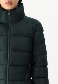Mujer vistiendo una chaqueta acolchada de color verde oscuro con cremallera y cuello alto frente a un fondo blanco liso.