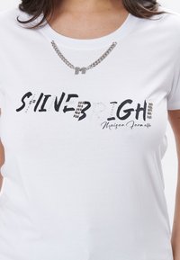 Hvid t-shirt med teksten "SHINE BRIGHT" i sort og sølv rhinsten, prydet med en sølv kæde med et "M" vedhæng.
