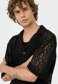 Camisa de encaje negro con un patrón de diamantes, de mangas cortas y cuello de botonadura. Presenta un diseño suave y texturizado y un corte relajado.